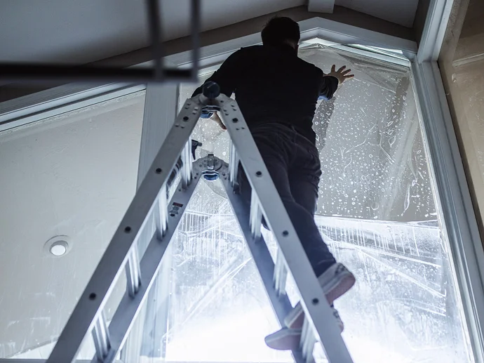 Person standing on a ladder inside a house applying protective or tinting film to a large triangular window with cleaning solution visible on the glass.