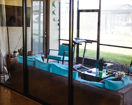 View through glass doors into a screened-in patio furnished with turquoise cushions on wicker furniture, a treadmill, and a small table with drinks and plants, overlooking a grassy backyard.