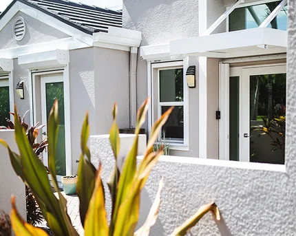 Exterior view of a modern light gray home with white trim, sliding glass doors, and potted plants, partially obscured by tropical foliage in the foreground.