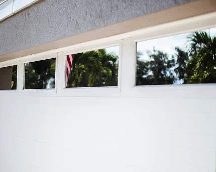 Close-up of a modern white garage door with four rectangular glass windows reflecting palm trees and part of an American flag.