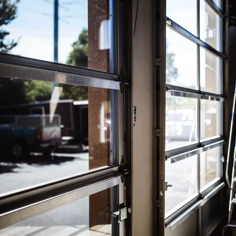 Close-up interior view of a modern glass-panel garage door partially opened, showing the metal tracks and outdoor light streaming through the transparent panels.