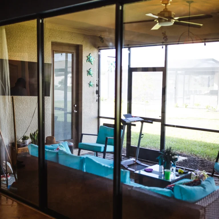 Enclosed patio viewed through glass doors, featuring wicker furniture with turquoise cushions, a treadmill, decorative wall turtles, ceiling fan, and a view of the grassy backyard.