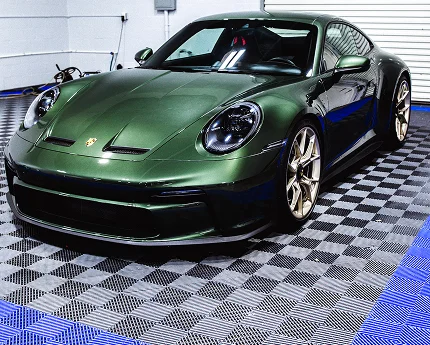 A dark green Porsche sports car with gold rims is parked indoors on a blue and gray checkered garage floor under bright lighting.