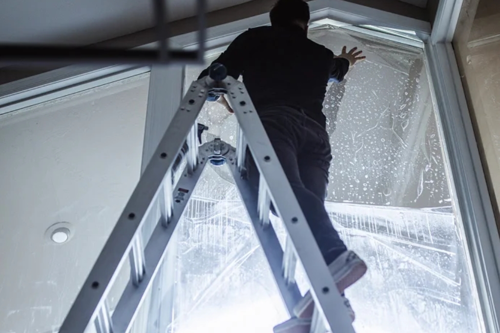 Technician on a ladder applying window film to a large upper-level residential window, illustrating the complexity and labor involved in estimating how much home window tinting costs based on size, access, and film type.