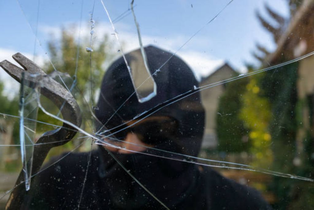 A masked intruder attempts to break a window with a crowbar, but the cracked glass remains intact due to security window film, illustrating the benefits of home window tinting for burglary resistance.