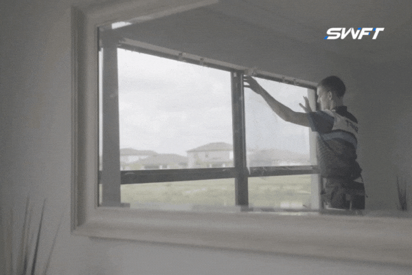 A window tinting specialist from Fort Myers Tint applies film to a large residential window, with the reflection showing surrounding homes in a sunny Fort Myers neighborhood. The process demonstrates the professional installation of protective architectural window film.