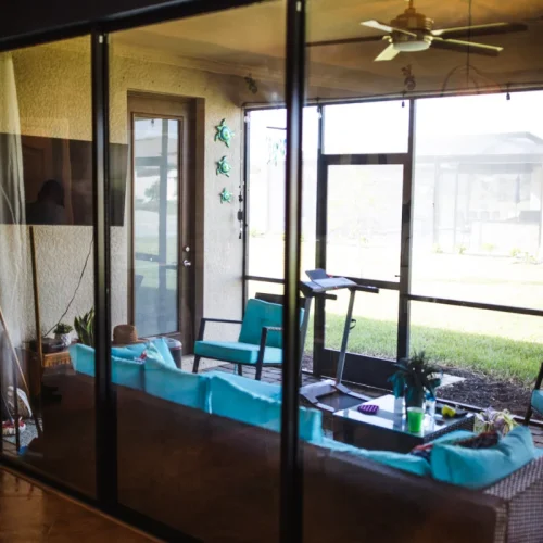A cozy enclosed patio is seen through large sliding glass doors, featuring turquoise cushioned seating, a treadmill, and decorative wall art. Natural light fills the space, with a view of the green lawn outside.