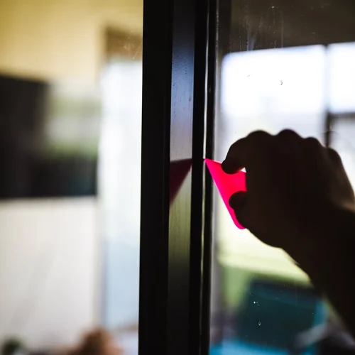 Person using a small pink squeegee to apply or smooth window film on a glass door, ensuring a clean and bubble-free finish. The scene is softly lit with a blurred background indoors.
