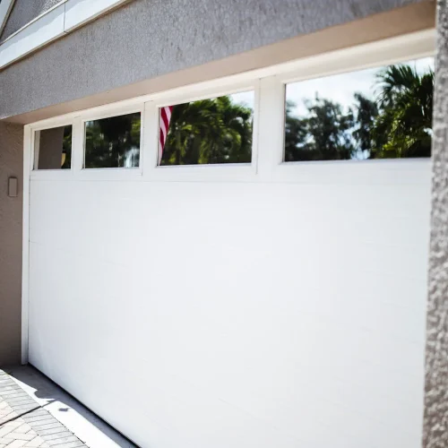 Modern white garage door with four rectangular glass windows reflecting palm trees and part of an American flag, set against a stucco exterior wall.